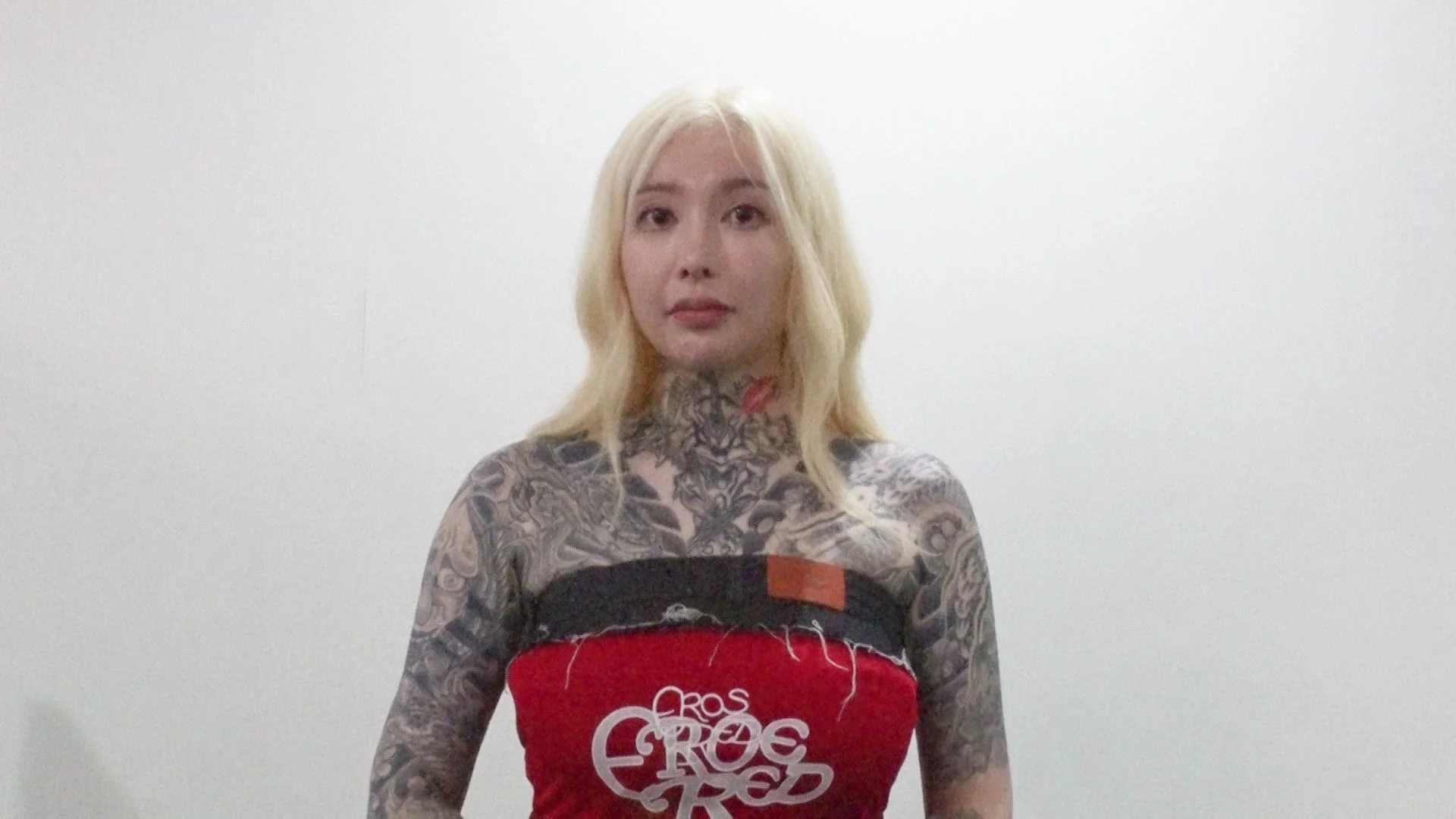 金髪で全身にタトゥーを入れた女性が赤いチューブトップを着用し、白い背景の前で正面を向いている